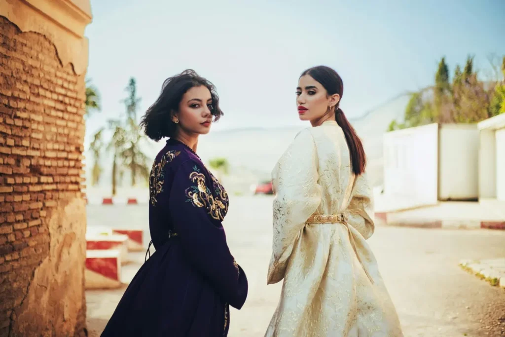 Two women wearing traditional Moroccan caftan and takchita with embroidered details, photographed outdoors in a sunlit street